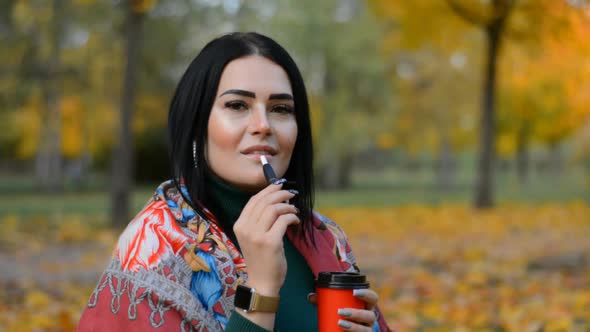 Portrait of a Beautiful Brunette Girl in an Autumn Park Drinks Coffee and Smokes an Electronic