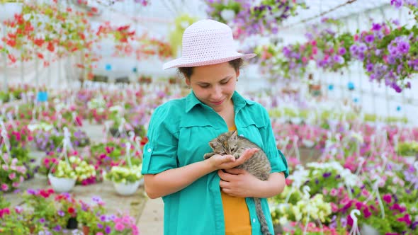 A Joyful Young Lady of a Greenhouse with Flowers Holds a Gray Kitten in Her Hands