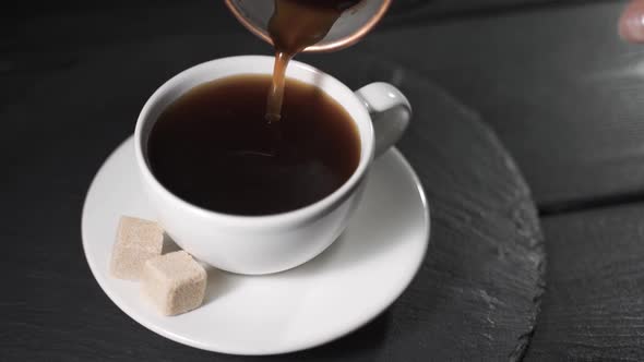 Pouring Coffee From Coffee Cezve (Turka) Into the White Cup on Dark Background