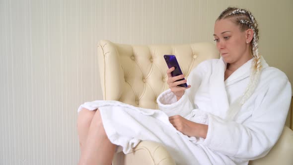 Young woman in white bathrobe uses mobile phone while sitting on armchair.