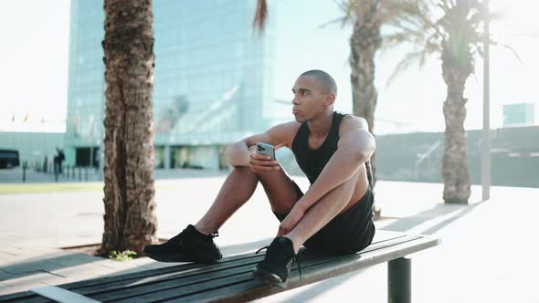 Bald meditative sportsman wearing black T-shirt typing by phone on the bench