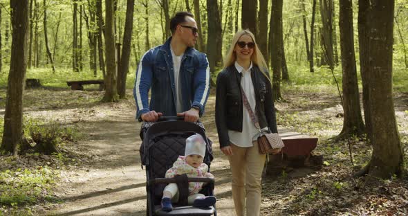 Happy Mom Dad With Baby In Stroller Walking In The Park Smiling