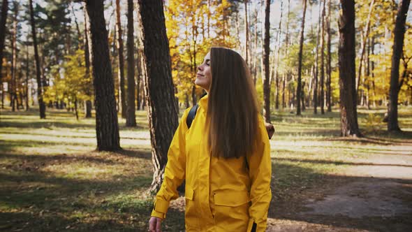 Young Lady in Parka Jacket and Backpack is Smiling While Walking By Path in Autumn Wood on Sunny Day