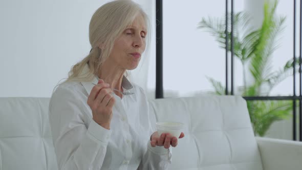 Elderly Whitehaired Woman Eats Yogurt Sitting on the Couch
