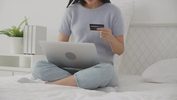 Young asian woman sitting on bed using laptop computer shopping online with credit card.