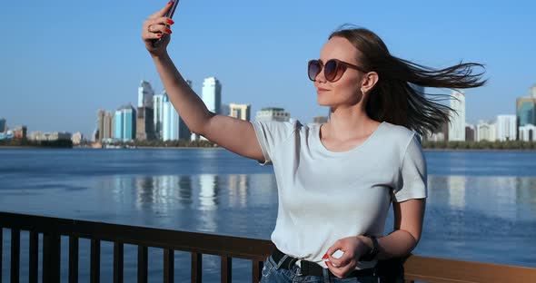 In Summer, the Girl on the Background of the Big City and the Waterfront Takes Selfie on Smartphone