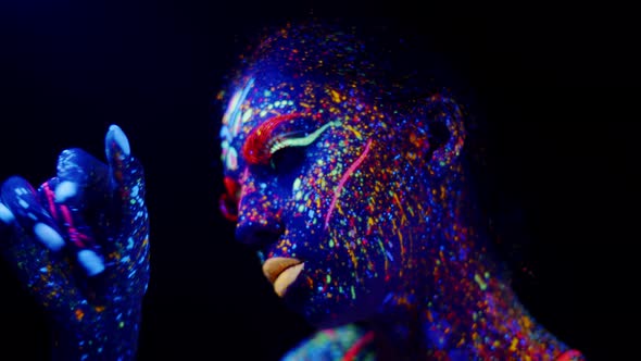 Woman in neon party makeup glowing in ultraviolet light touching her face, looking at her hand.