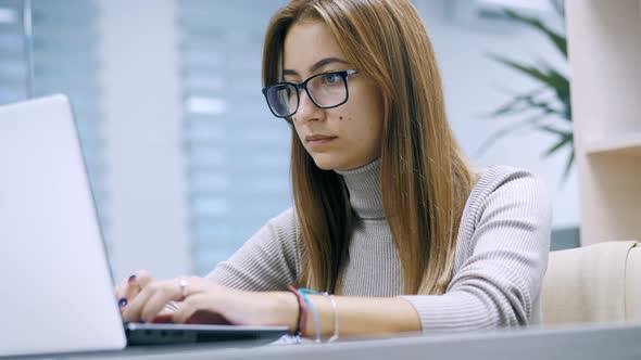 Cute Woman Working at a Laptop with Glasses Typing Text in the Office Employee at Work on the