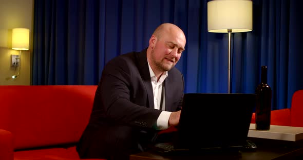 Adult Bald Man Dressed in Black Suit Is Drinking Wine and Calling By Video By Notebook in Living