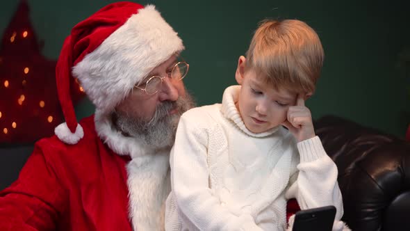 Santa Claus with a Child Sitting on His Lap are Taking a Selfie Using a Smartphone
