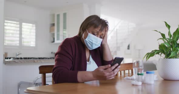 African american senior woman in face mask using smartphone at home, with medication on table