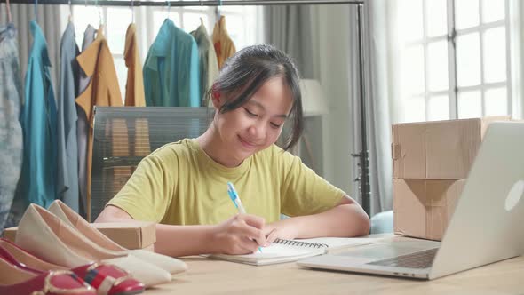 Young Girl Online Seller Looking At Computer And Writing In The Notebook While Selling Clothes