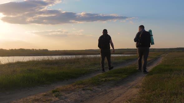 Two Friend Tourists Walk Along the Lake at Sunset, Traveling with Backpacks in Warm Weather 