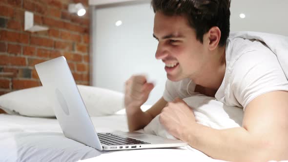 Young Man in Bed Working on Laptop and Reacting to Success