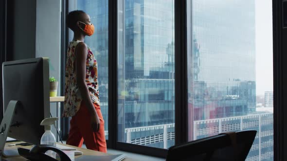 Thoughtful african american woman wearing face mask looking out of window at modern office