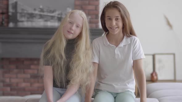 Two Cute Smiling Girls Sitting on the Couch Ooking at Camera in the Living Room. Concept of