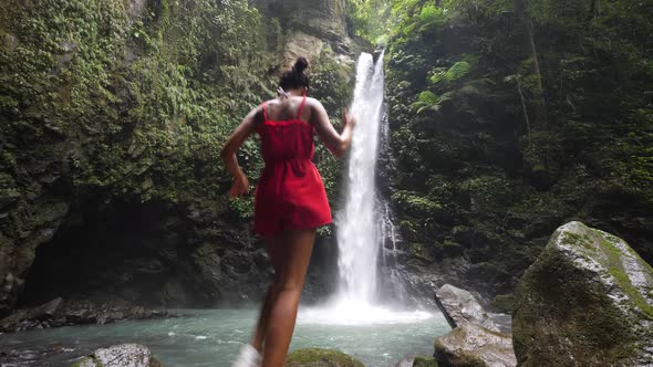 Young Woman in a Red Romper Walks and Jumps at the Foot of a Waterfall