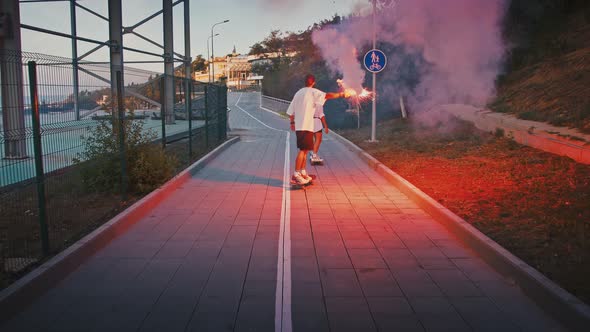 Active Young Couple in Love Riding Longboards with Red Signal Fire in Evening