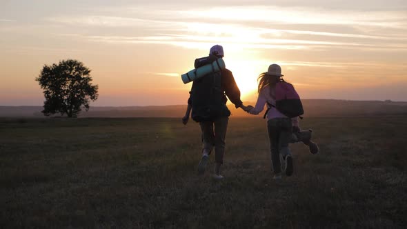 Mother and Daughter with Backpacks Running Through the Meadow at Sunset. Family Tourism Concept.