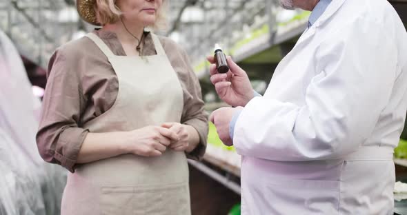 Unrecognizable Caucasian Man Giving Bottle with Fertilizer To Mature Woman in Apron, Professional