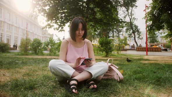Young Asian woman sitting on grass in the park holding smartphone, using mobile apps. Stylish girl.