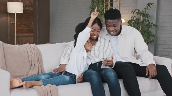 African Family Parents with Kid Child Girl Daughter Relax on Sofa Home Living Room Using Smartphone