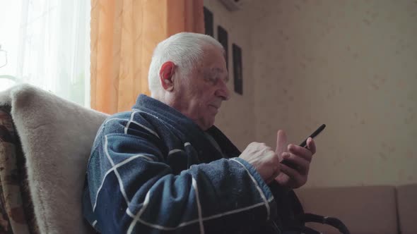 Elderly Man with Smartphone Sitting in an Armchair and Scrolling Social Media