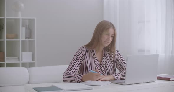 Female Student Is Learning Online, Viewing Video Lesson on Laptop and Writing in Exercise Book