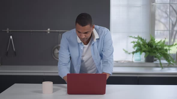 Young Handsome Concentrated Man Looking at Laptop Screen Standing in Kitchen