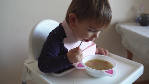 Young Toddler Girl Witth Silicone Bib Eating Alone Soup with Spoon