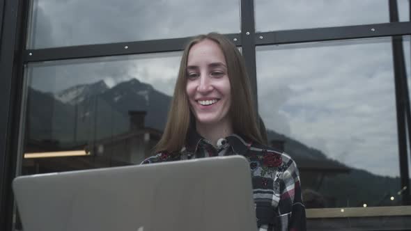 A Young Beautiful Girl Uses a Laptop for a Video Call in a Cafe Against the Backdrop of Mountains