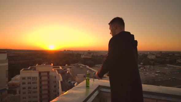 Close the Lid of the Laptop and Drink Beer Standing on the Roof. A Man in a Hoodie with a Laptop in