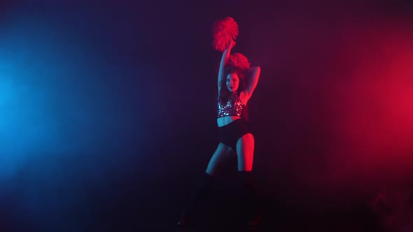 Young Cheerleader with Pompoms in Uniform is Dancing on Black Smoky Studio Background with Red and
