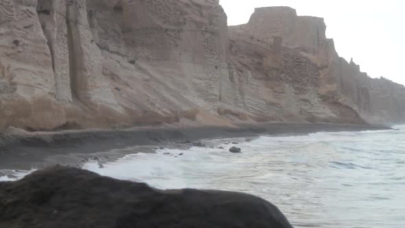 Tracking shot of waves hitting a black beach that's surrounded by tall white volcanic cliff formatio