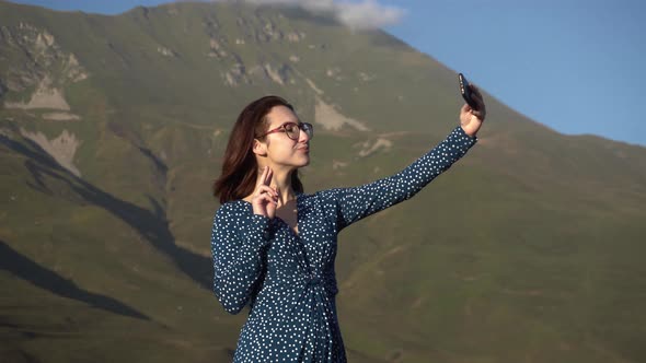 A Young Woman in a Dress Stands in the Mountains and Takes a Selfie on a Smartphone.