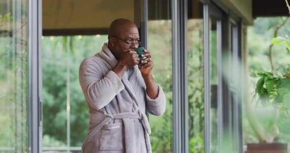 African american senior man standing on balcony wearing bathrobe drinking coffee and enjoying view