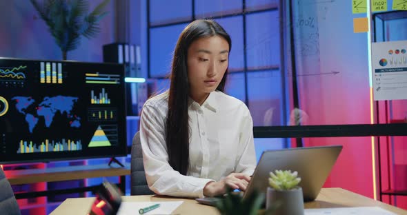 Asian Woman Sitting in front of Computer and Typing on It, Working Overtime in Modern Office