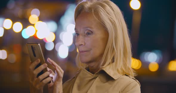 Bokeh Shot of Smiling Aged Lady Standing on Street at Night Replying to Text on Cellphone