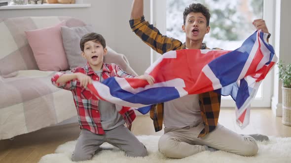 Happy Middle Eastern Teenager and Boy Rejoicing Soccer Team Victory Shaking British Flag Watching TV