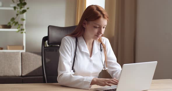 Serious Red Haired Girl Young Woman Doctor or Nurse Sits in Clinic Office with Laptop Works Online