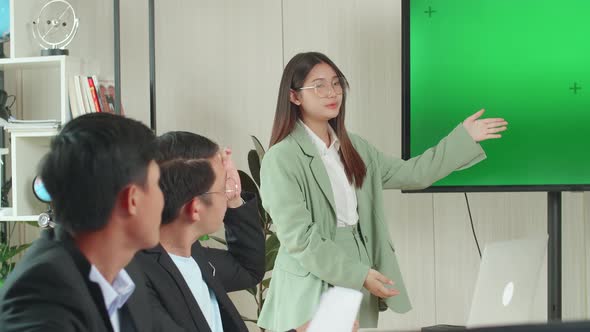 Asian Businesswoman Presentation To Her Business Colleagues, Pointing At Tv Mock Up Green Screen