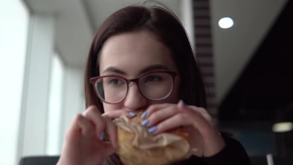 A Young Woman Is Eating a Burger. A Girl Sits in a Cafe By the Panoramic Window and Eats Fast Food.