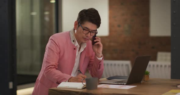 Smartly dressed asian businessman sitting at desk using laptop taking notes