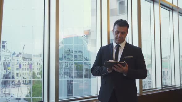 The Satisfied Man Is Standing at the Window and Uses His Tablet