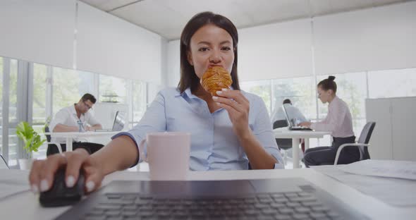 Latin Businesswoman Sitting at Desk with Coffee Using Laptop and Eating Croissant