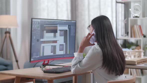 Asian Woman Engineer Talking On Smartphone While Designing House On A Desktop At Home