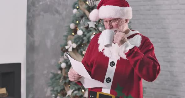 Smiling Old Caucasian Man in Santa Claus Costume Drinking Coffee and Reading Letter