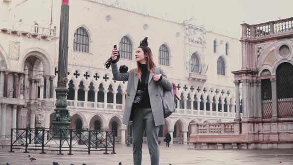 Happy Smiling Female Tourist with Pigeons Sitting on Her Arm and Head Takes Selfie on City Square in