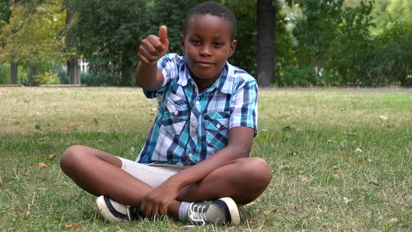 A Young  Black Boy Sits on Grass in a Park and Shows a Thumb Up To the Camera with a Smile