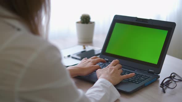 A nurse in a bathrobe is typing on a laptop with a green screen on the table